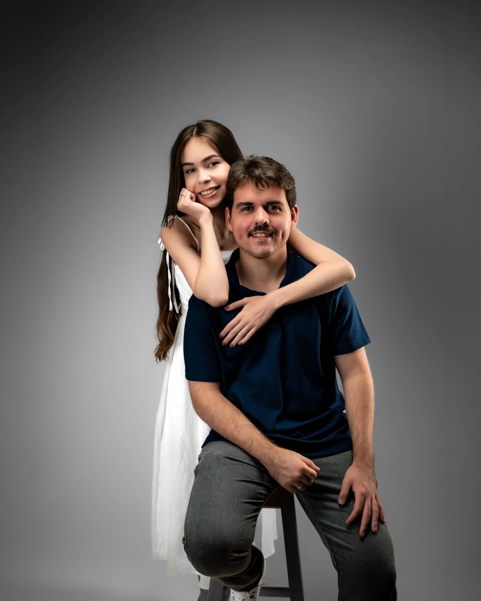 Ele sentado no banco, ela apoiada em seus ombros, ambos olhando para a câmera