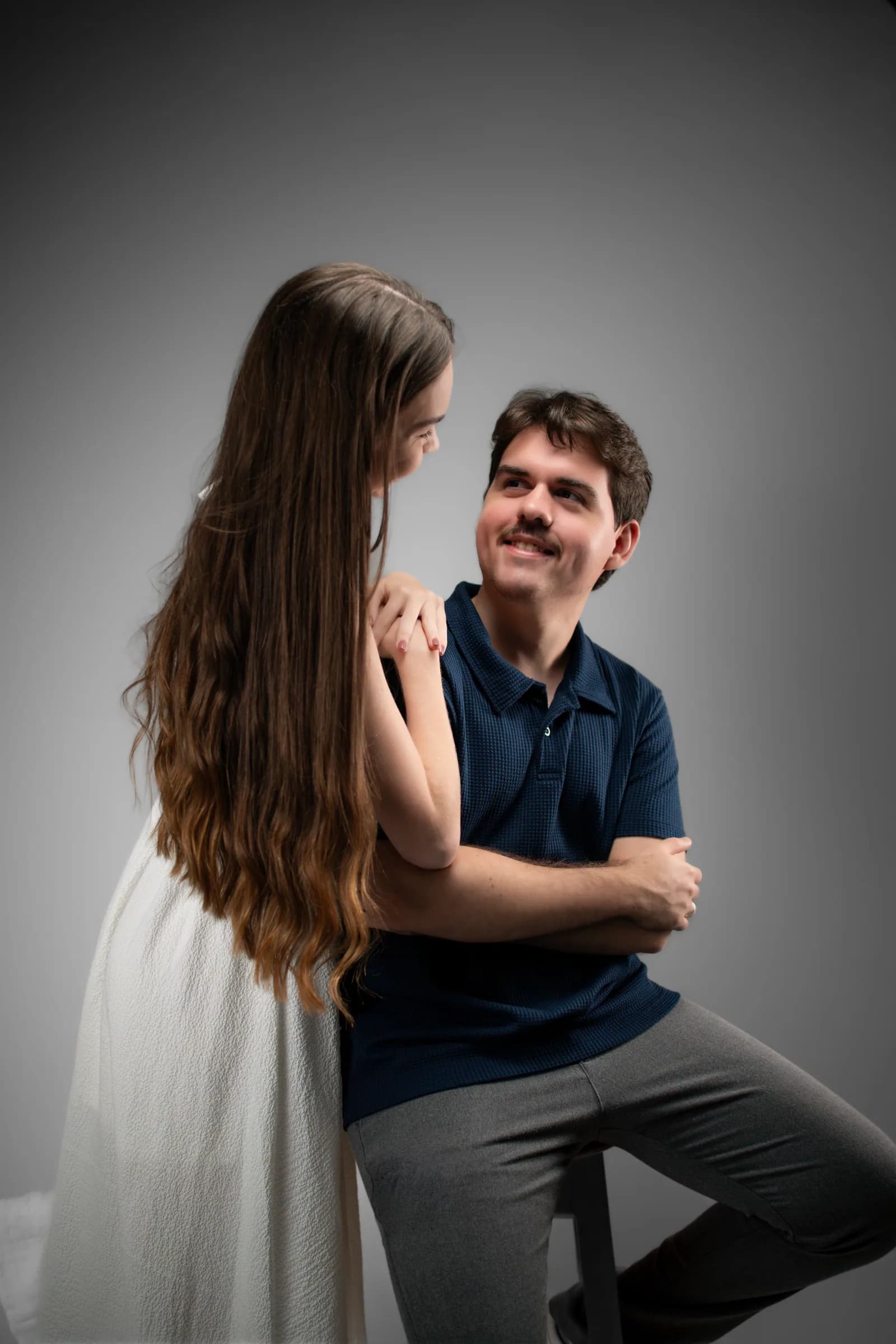 Ele sentado olha para ela, de pé e de costas para a câmera
