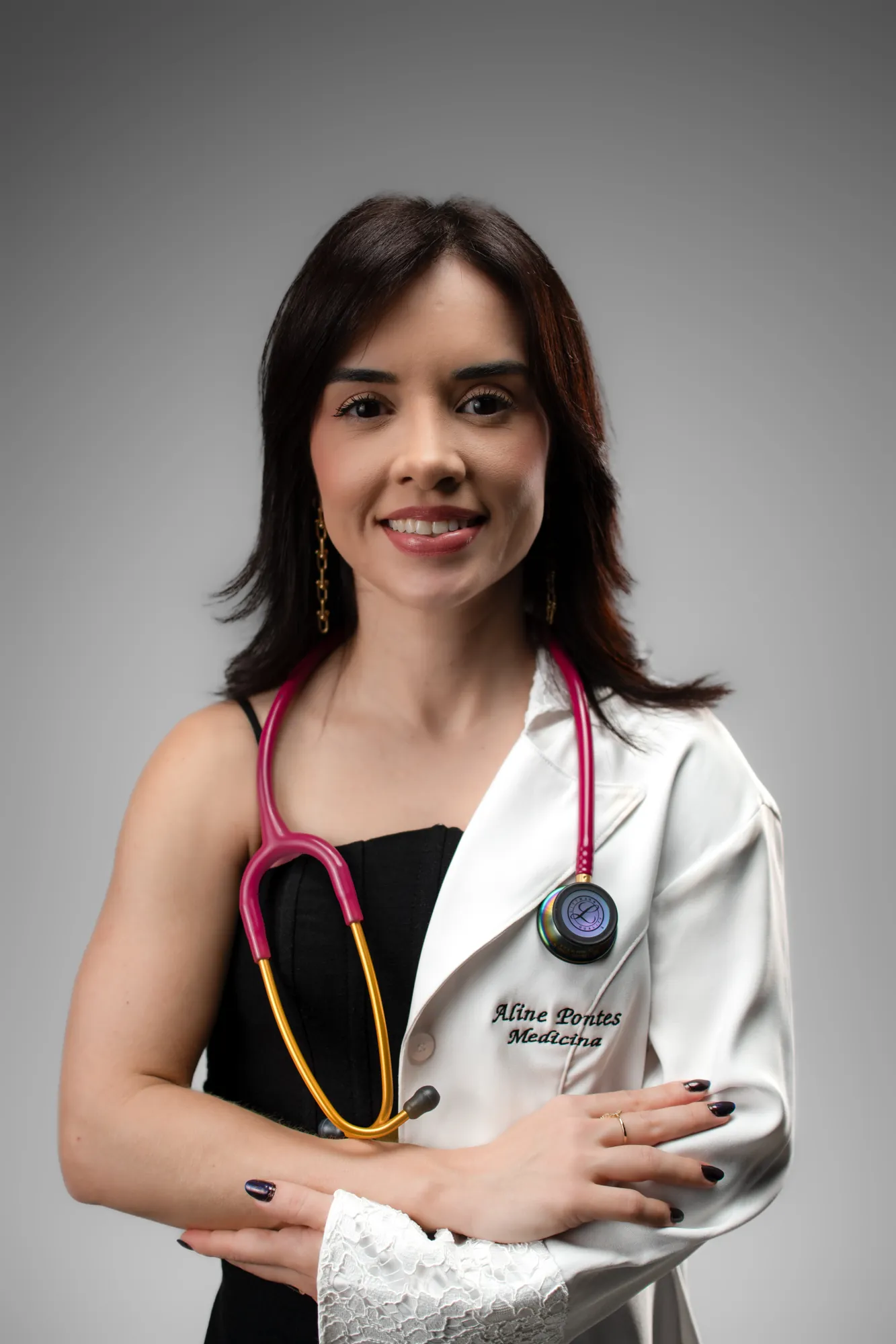 Médica de jaleco branco e estetoscópio rosa, braços cruzados, sorrindo para a câmera