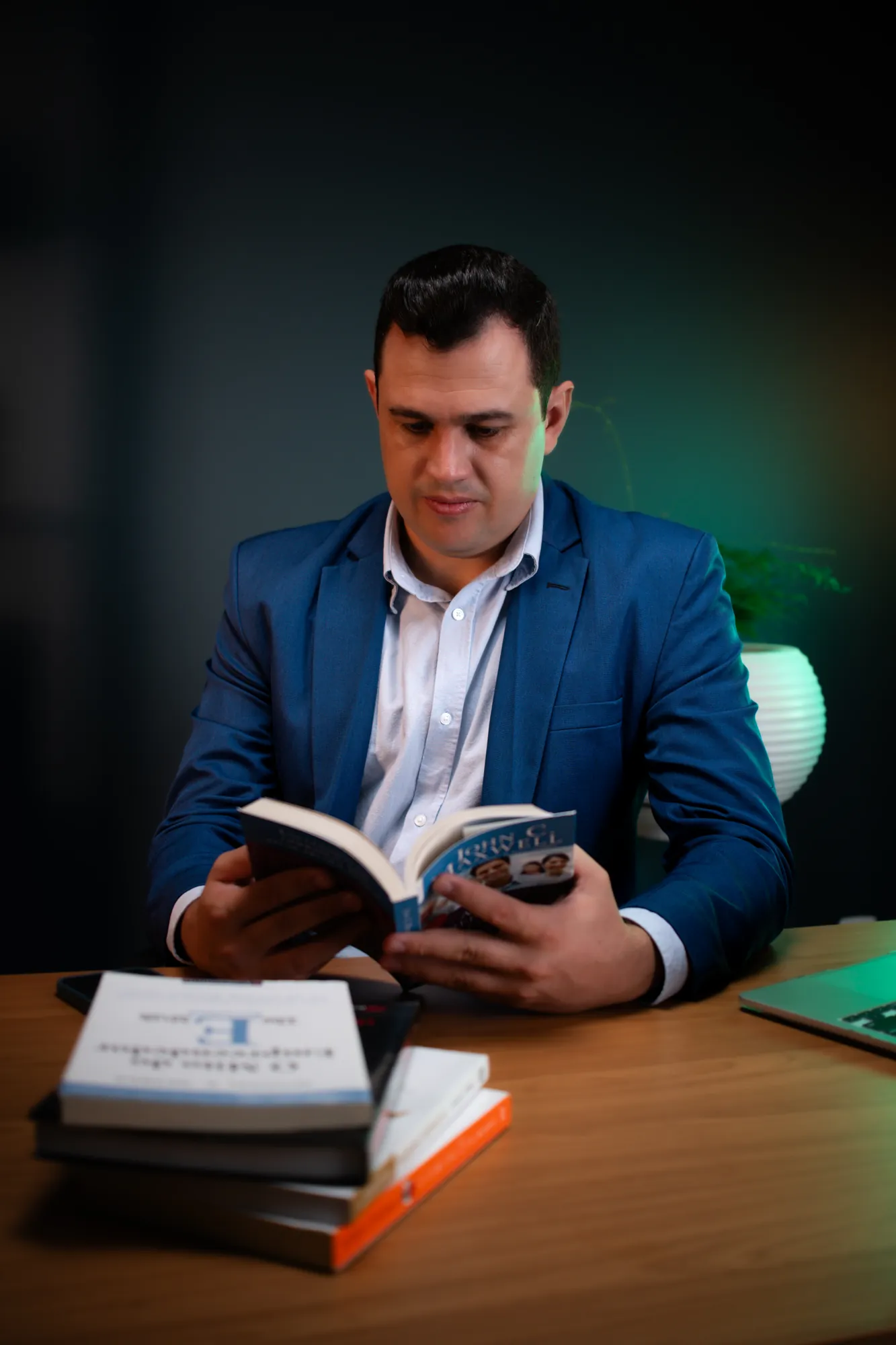 Homem de terno azul lendo um livro à mesa de escritório, com luz verde ao fundo