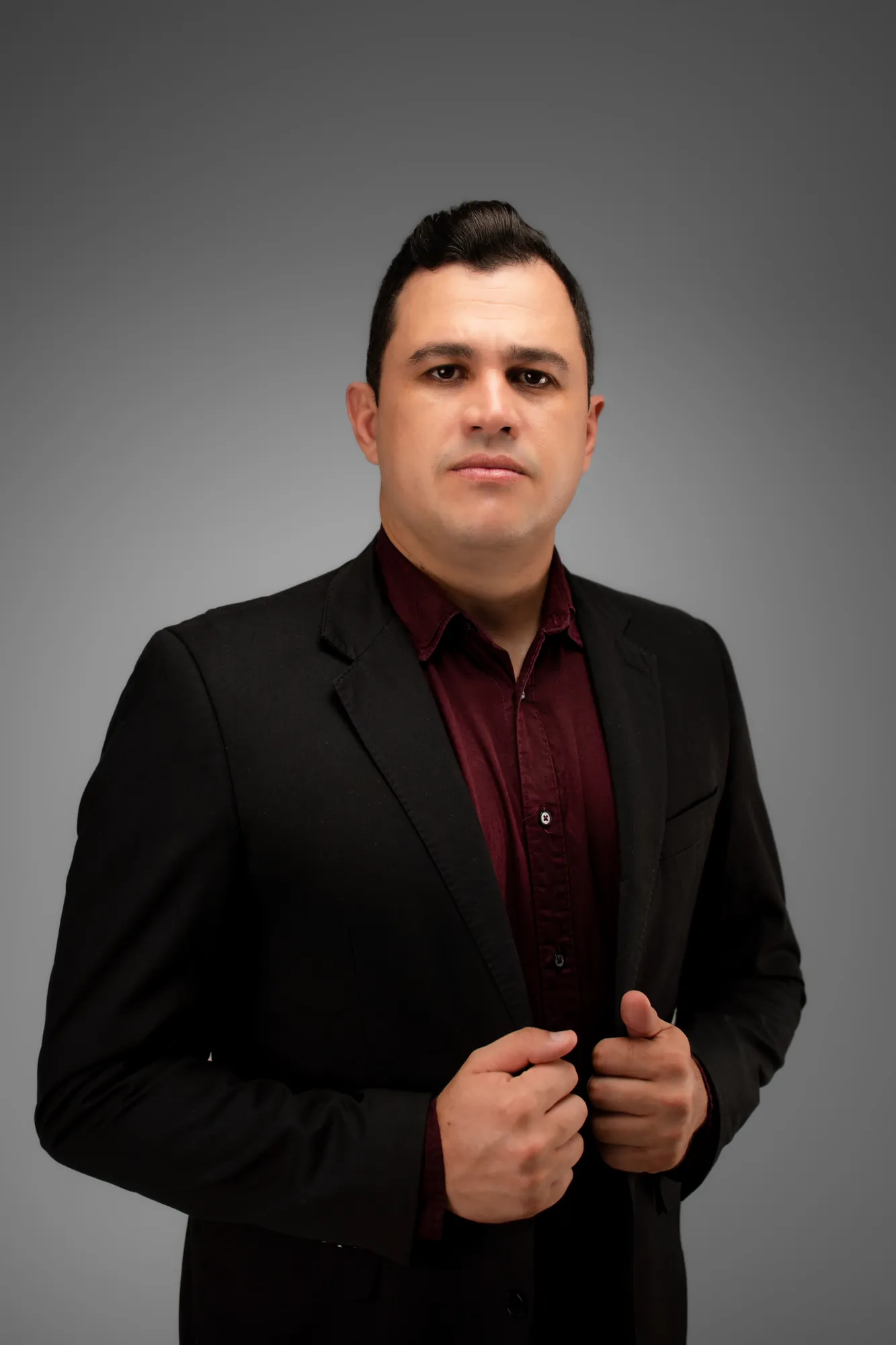 Homem de terno preto e camisa vinho, de frente, ajeitando o paletó