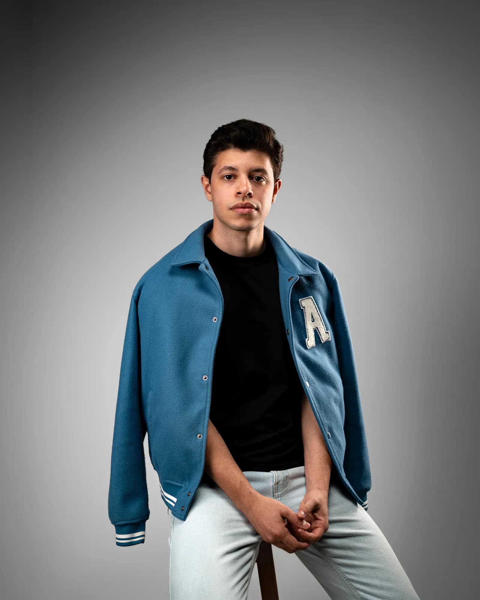 Jovem de jaqueta azul de time abrindo a gola, sentado numa banqueta