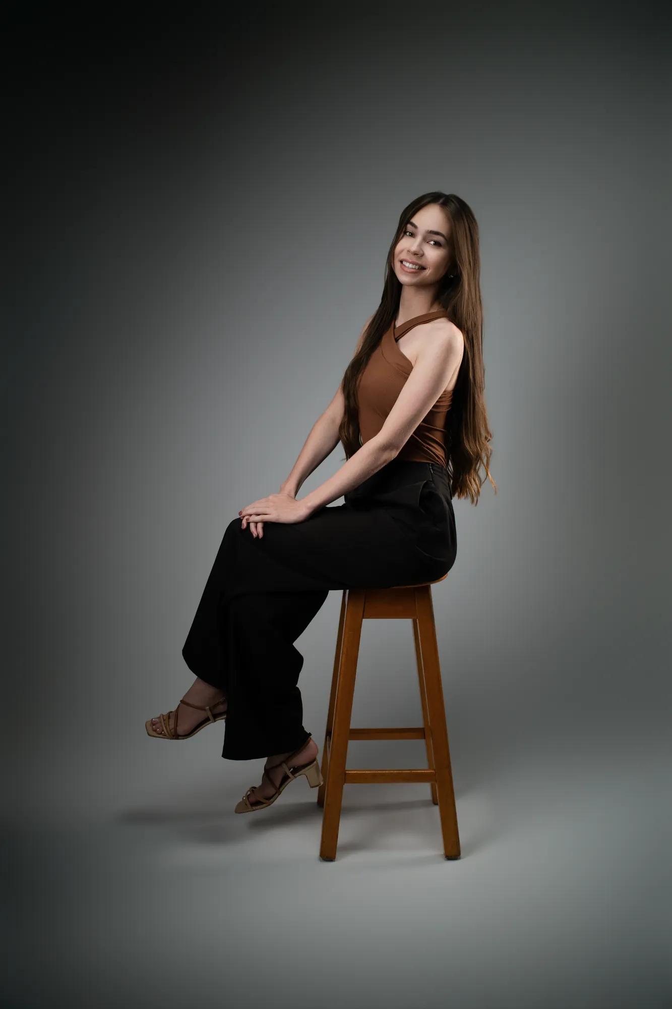 Mulher de blusa marrom e calça preta sentada numa banqueta de madeira, olhando para a câmera