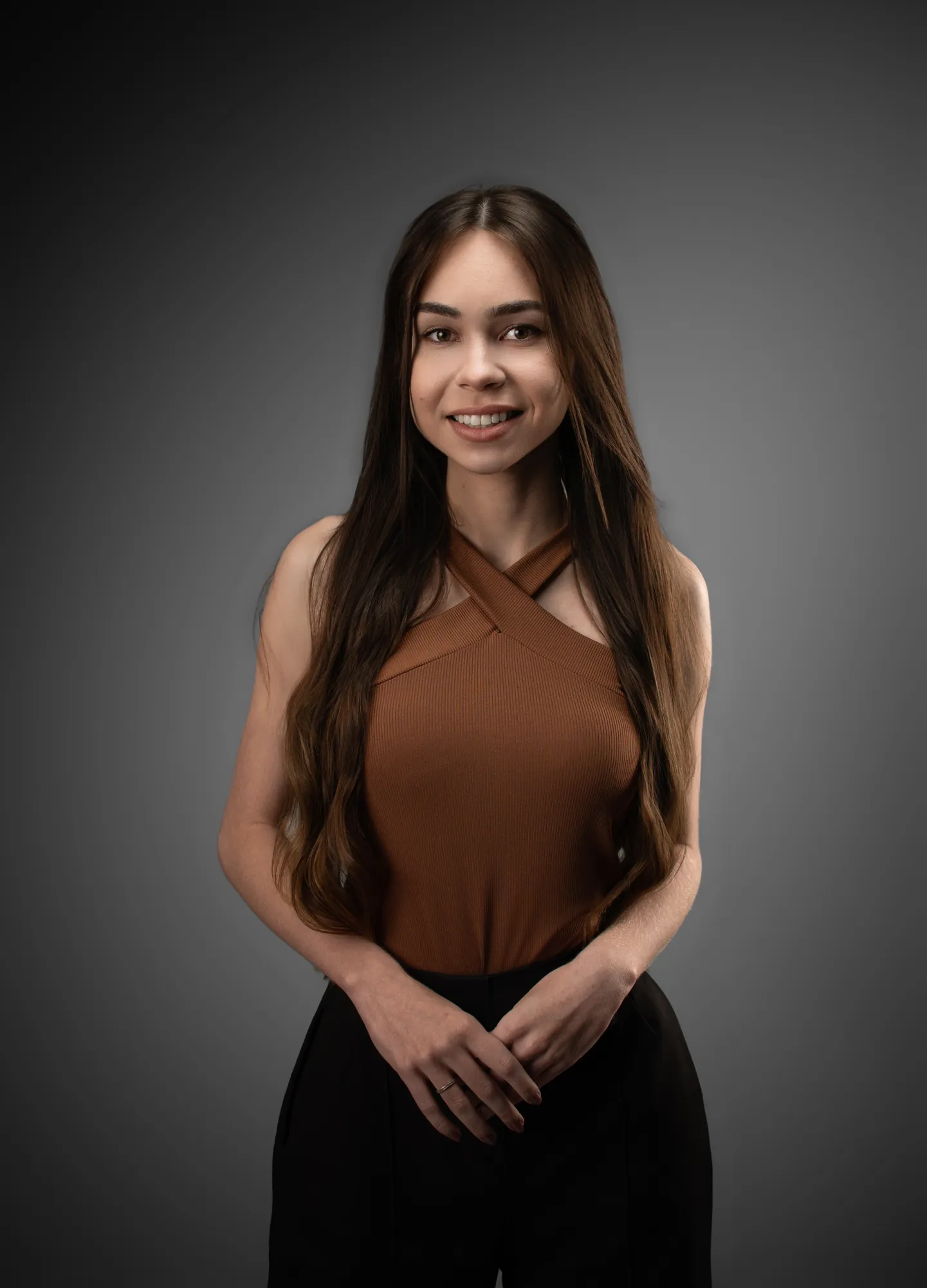 Mulher de corpo inteiro com os braços abertos, blusa marrom e calça preta, em fundo cinza